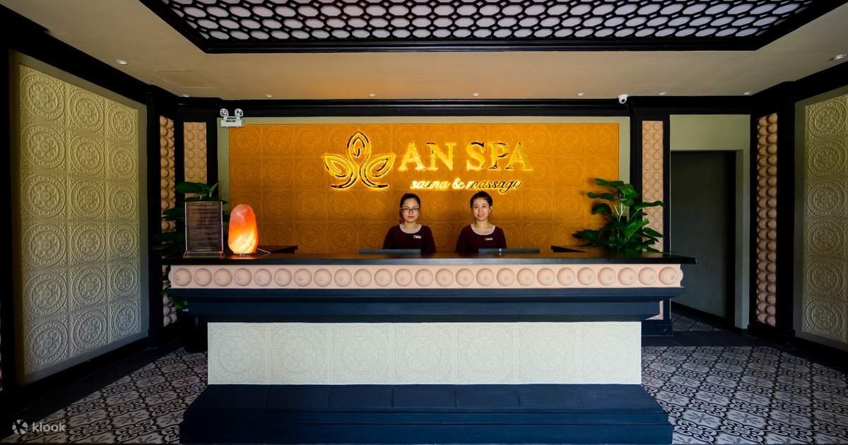 Quầy lễ tân spa với hai nhân viên đứng phía sau, nền tường trang trí và cây xanh bên cạnh.