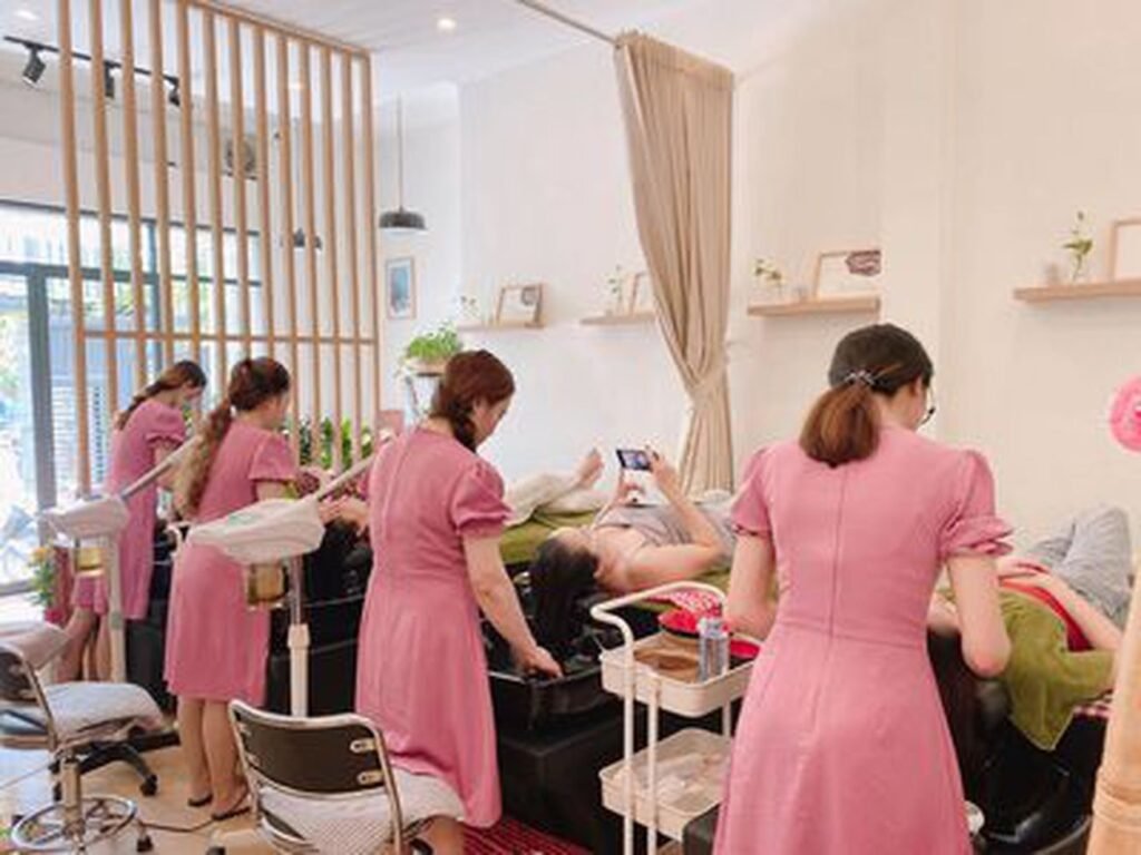 Nhân viên mặc đồng phục hồng chăm sóc khách hàng tại một tiệm spa, với các thiết bị làm đẹp xung quanh.
