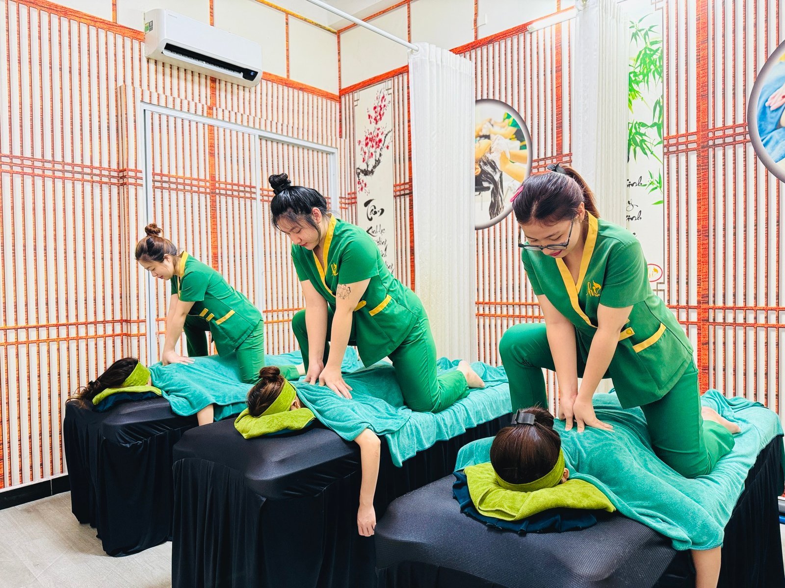 Nhân viên spa mặc đồng phục xanh đang thực hiện massage cho khách trên giường có phủ khăn màu xanh.