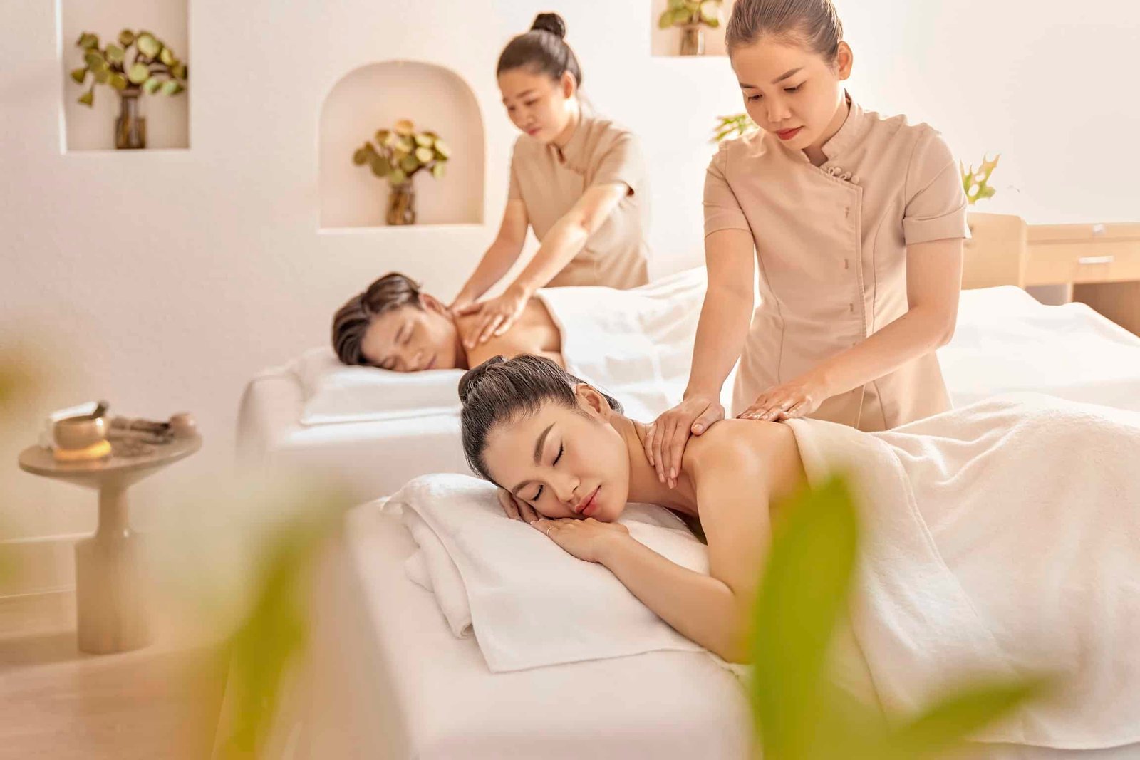 Hai người đang được mát-xa thư giãn trong phòng spa, có nhân viên phục vụ và không gian ấm cúng.