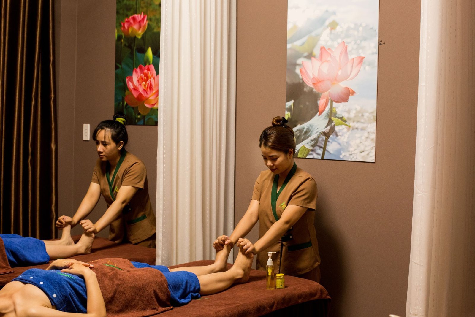 Hai nhân viên spa đang thực hiện massage chân cho khách hàng trong phòng có tranh hoa sen.