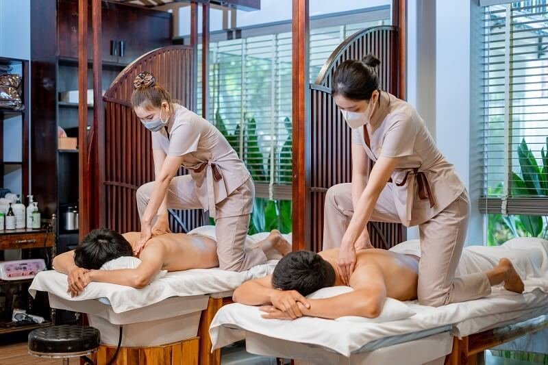 Hai nhân viên đang thực hiện massage cho hai người nằm trên giường trong một không gian spa thư giãn.