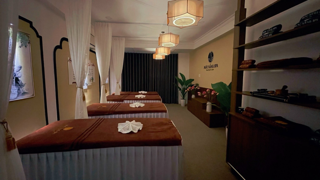 Phòng spa với ba giường mát-xa, rèm trắng, ánh sáng ấm áp và kệ chứa khăn tắm.