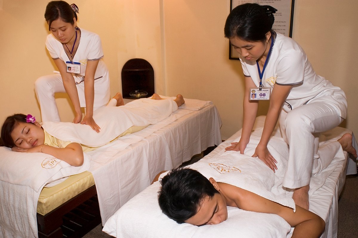 Hai người đang được mát-xa trong phòng spa, mỗi người nằm trên một giường riêng biệt có khăn trắng.