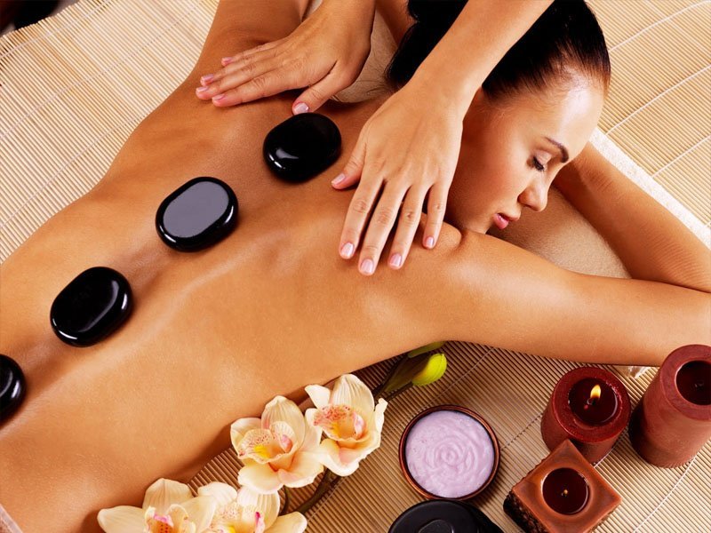 Người đang nằm thư giãn với đá nóng trên lưng, xung quanh là nến, hoa và kem dưỡng da.