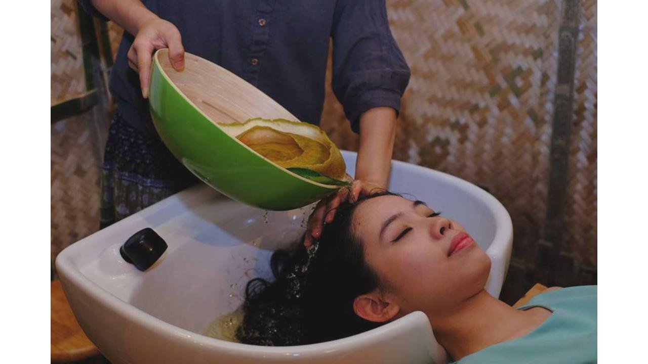 Người đang được gội đầu trong bồn rửa, nước được đổ từ tô xanh, không gian nền bằng tre.