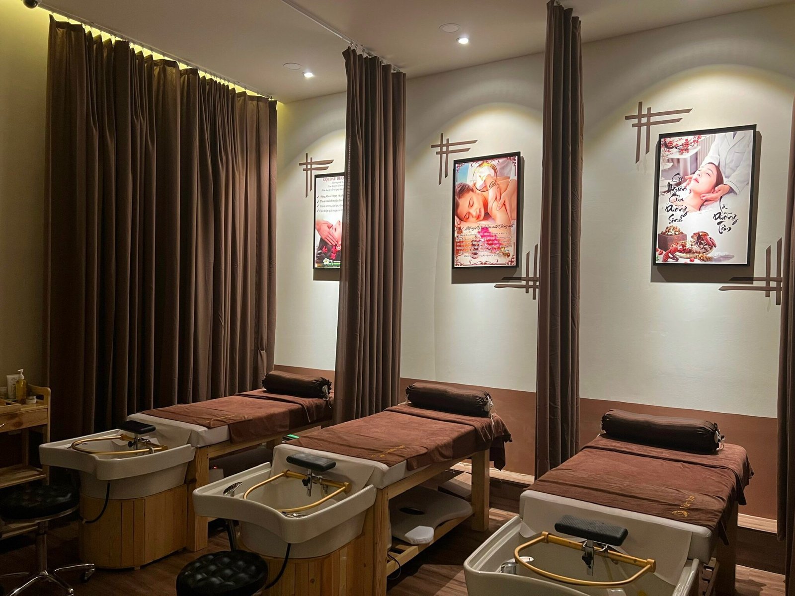 Phòng spa với giường massage, bồn rửa chân và tranh trang trí trên tường, rèm nâu tạo không gian thư giãn.