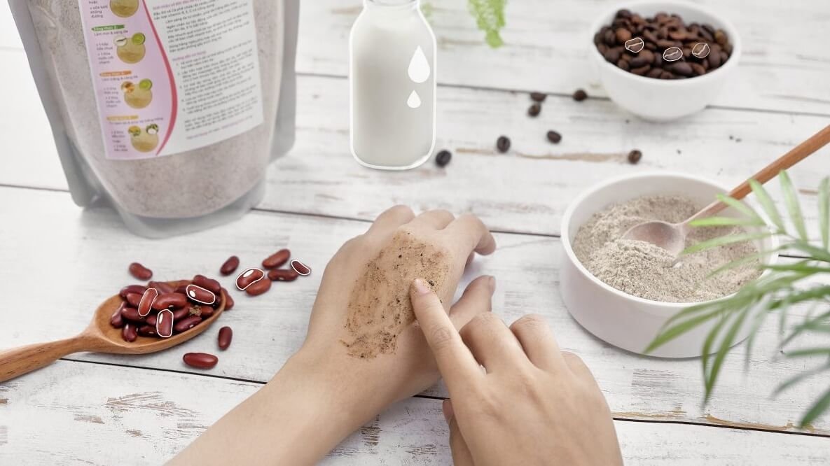 Người đang thoa bột lên tay, bên cạnh là bát bột, thìa gỗ đựng đậu đỏ, và ly sữa trên bàn gỗ trắng.