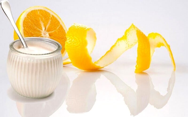 Hũ sữa chua đặt cạnh lát cam và vỏ cam xoắn, trên nền trắng sáng.